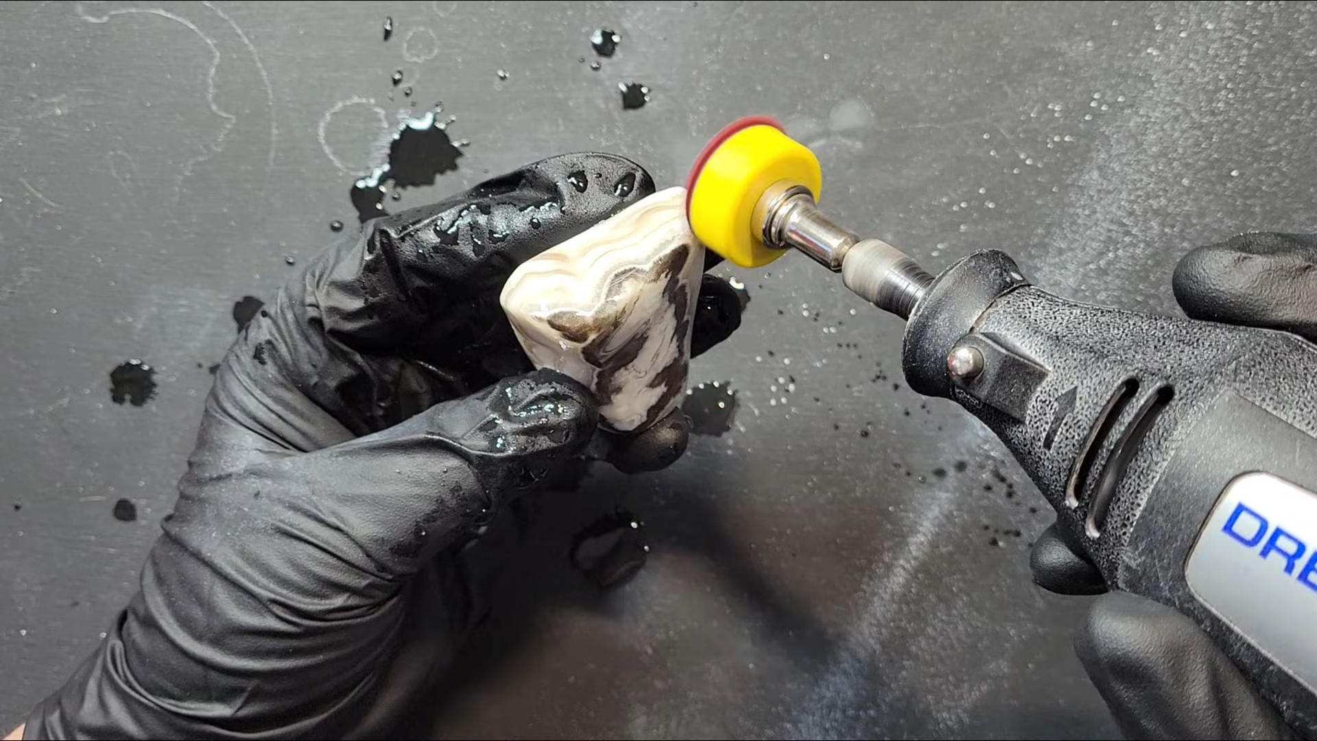 A Dremel rotary tool with polishing wheel attachment working on a banded calcite stone, held in gloved hands