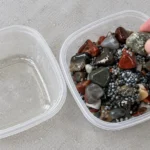 Hand selecting a polished tumbled stone from a container of mixed stones and ceramic media beside an empty rinse container
